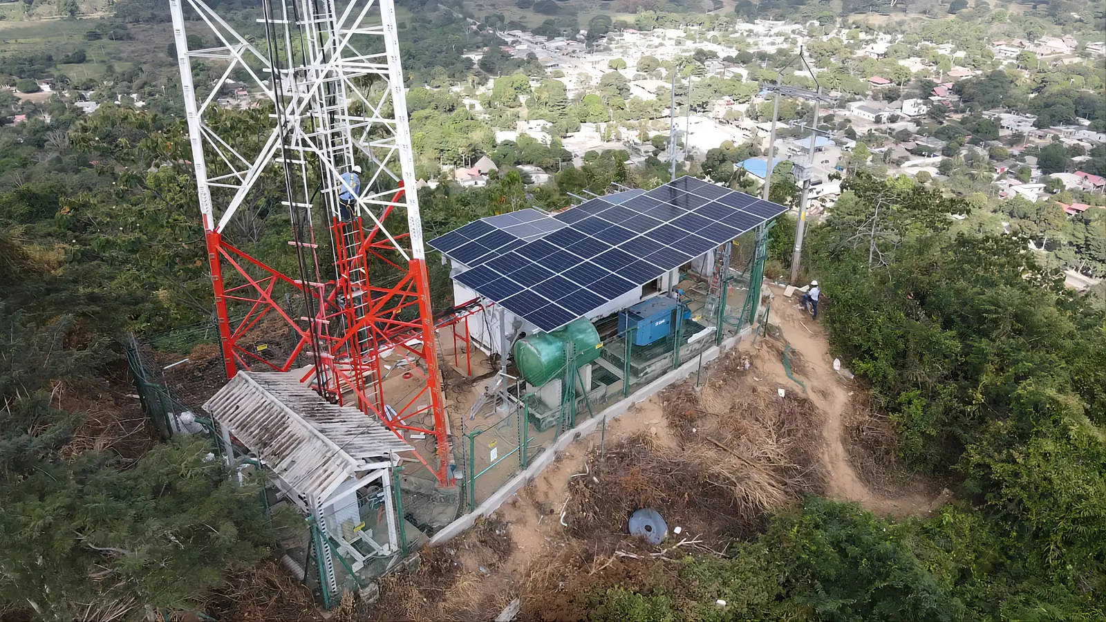 Vista aérea de una torre de telecomunicaciones en un cerro con arreglo solar, banco de baterías y cerramiento perimetral.